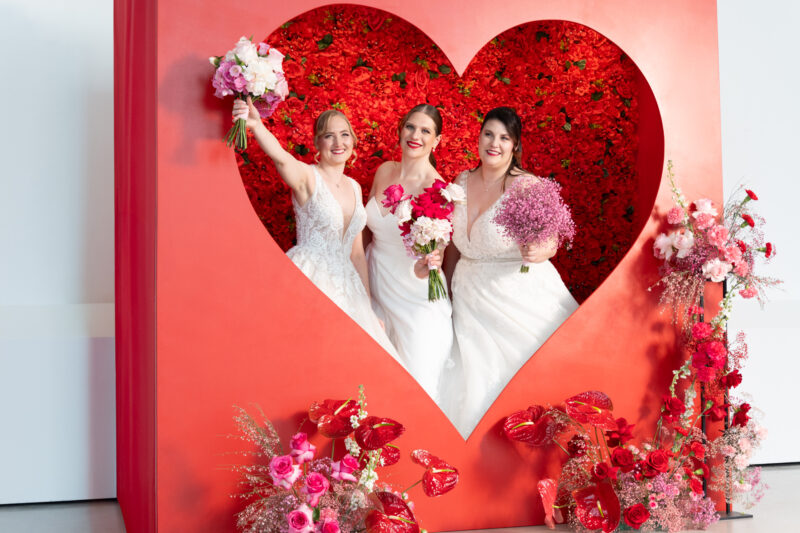 Bräute in der Fotospot Herz-Wall. Rote Flowerwall mit riesigem Herz